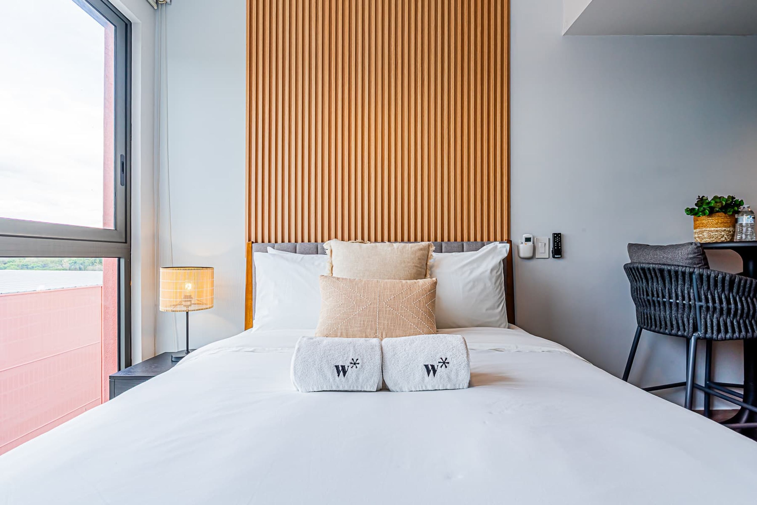 A neatly made bed with a headboard, pillows, and towels, a bedside lamp, and a chair with a small table beside the bed.