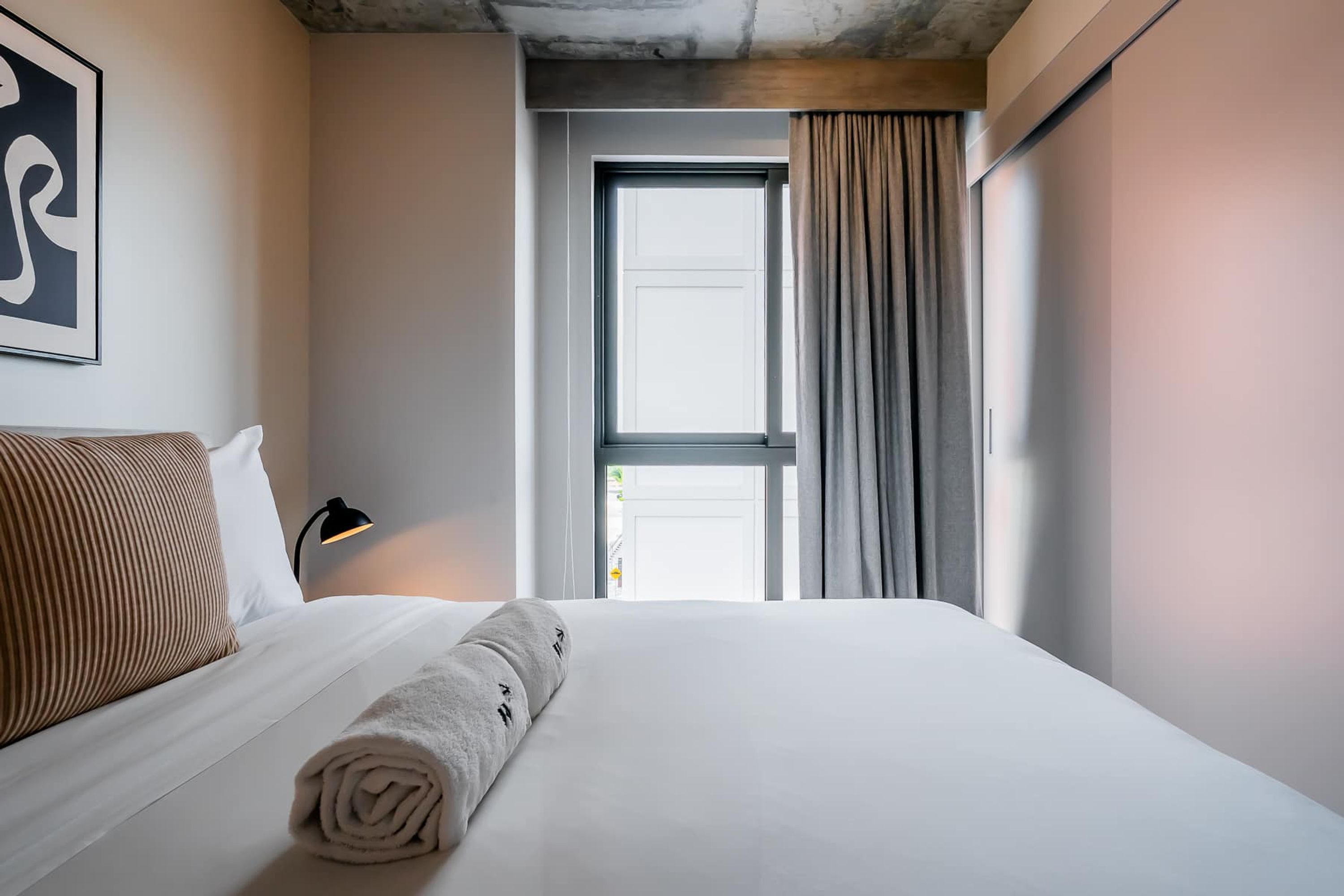 A hotel room with a bed, a window with curtains, a wall-mounted lamp, and a framed picture.
