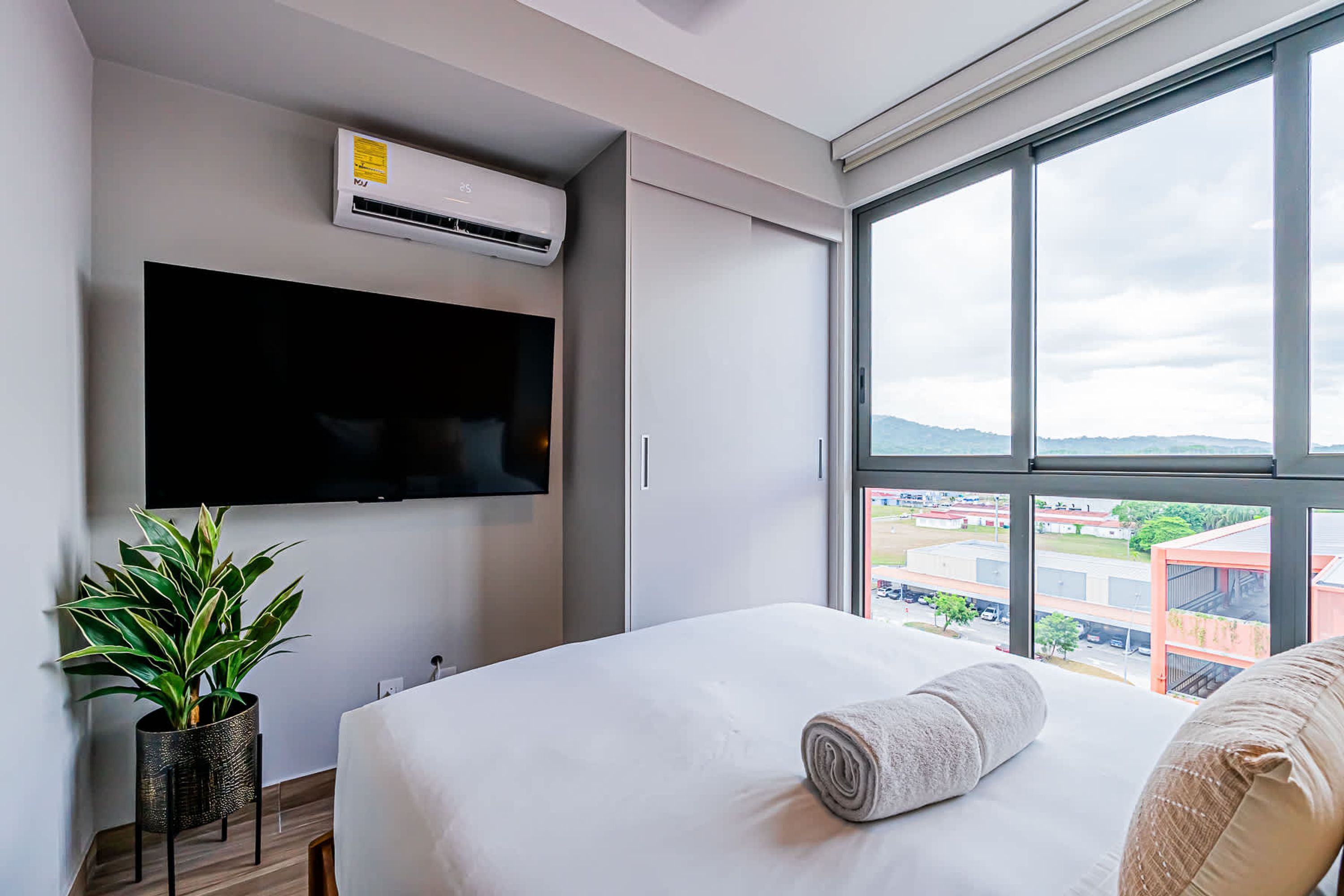 Une chambre d’hôtel moderne avec un grand lit, un téléviseur à écran plat fixé au mur, une plante en pot et une fenêtre donnant sur la ville.