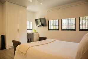 Habitación de hotel con una cama grande, un escritorio con silla, un televisor montado en la pared y ventanas con vistas a edificios.