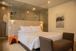 Habitación de hotel moderna con una cama grande, cabecera de madera, una mesita de noche naranja pequeña, un sillón marrón, un espejo grande y un cuadro en la pared.