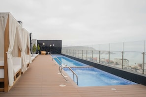 Una piscina en la azotea con vistas a la ciudad, terraza de madera y sillones de descanso.