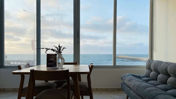 Une pièce comprenant une table à manger et des chaises, un canapé gris et de grandes fenêtres donnant sur la mer.