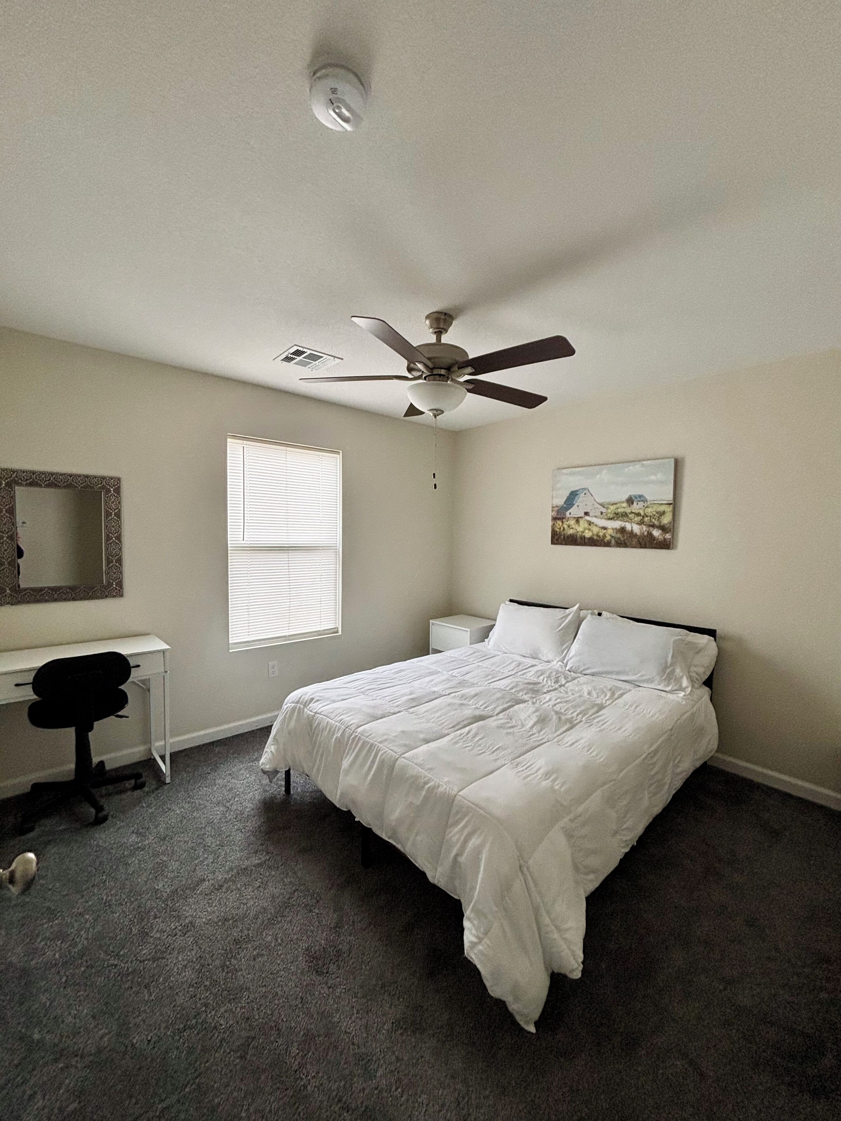 Una camera da letto con un letto, una scrivania, una sedia, una ventola a soffitto, una finestra e un quadro appeso al muro.