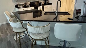 Une cuisine moderne avec des armoires blanches, un comptoir noir et des appareils en acier inoxydable. On y trouve deux tabourets de bar avec des assises rembourrées blanches et des structures métalliques.