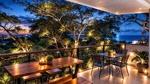 An outdoor dining area with tables and chairs, surrounded by trees and overlooking a coastal view.