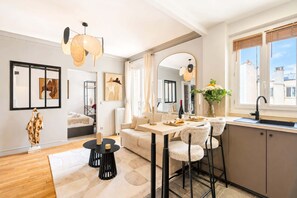 Una sala de estar moderna con sofá, mesa de comedor y sillas, un área de cocina con lavamanos y ventana, y obras de arte en las paredes.