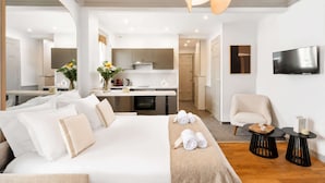 A modern hotel room with a large bed, a sitting area with a chair, a small table, a flat-screen TV mounted on the wall, and a kitchenette in the background. - Elegant apartment with lift - Gare de Lyon & Bercy (Paris)