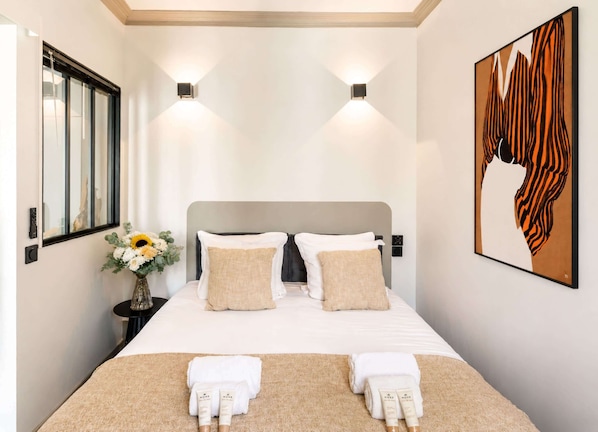 A hotel room with a bed, two wall-mounted lights, a painting, a vase with flowers, and a nightstand.