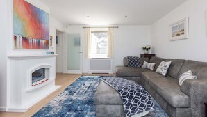 Een moderne woonkamer met een open haard, een grijze bank, een blauw tapijt en een kleurrijk abstract schilderij.