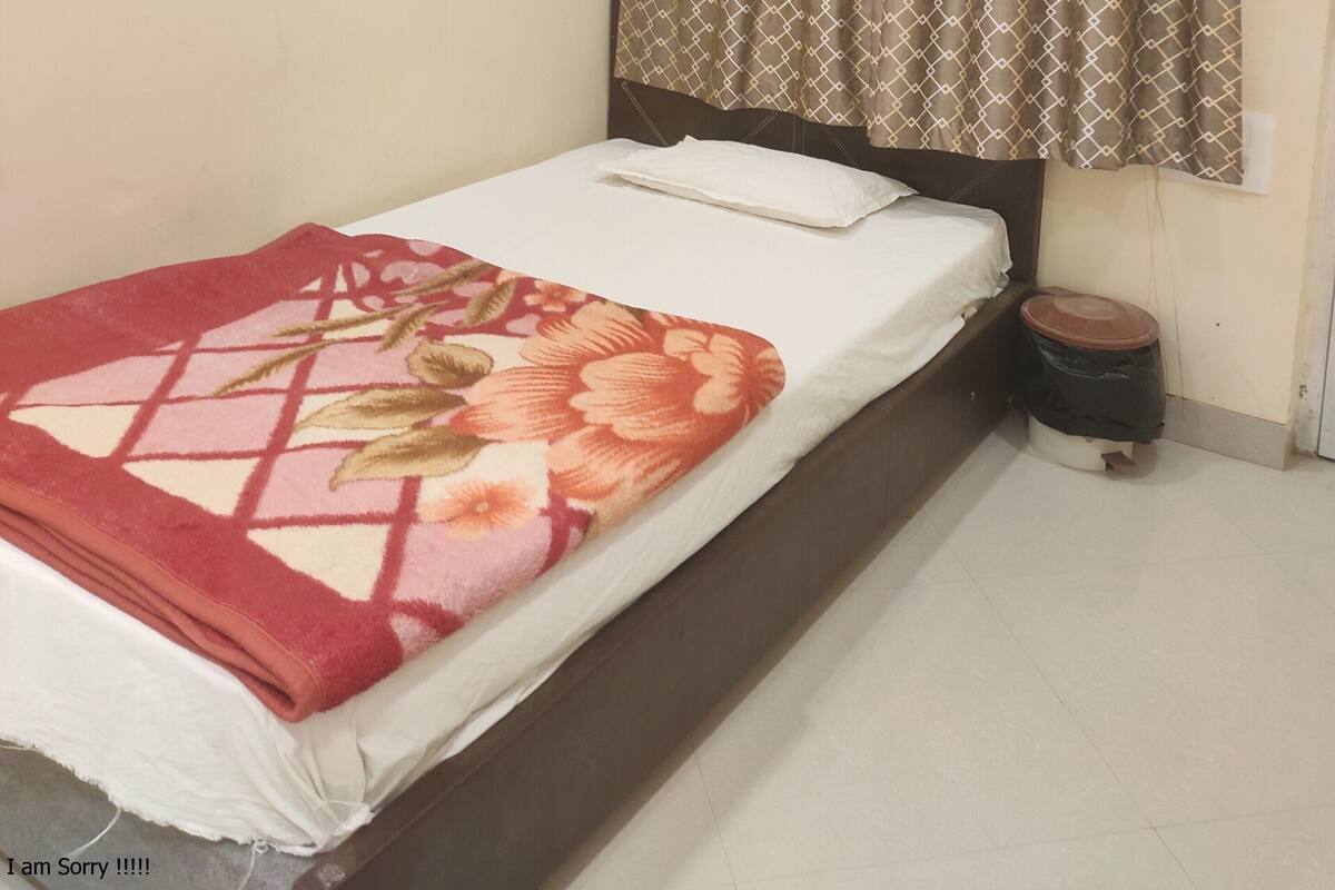 A single bed with a floral-patterned blanket, a bedside table with a trash bin, and an air conditioning unit on the wall.