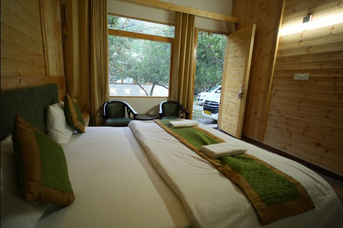 Una habitación con una cama, dos sillas, un ventanal con vistas a árboles y coches aparcados, y paredes de madera.