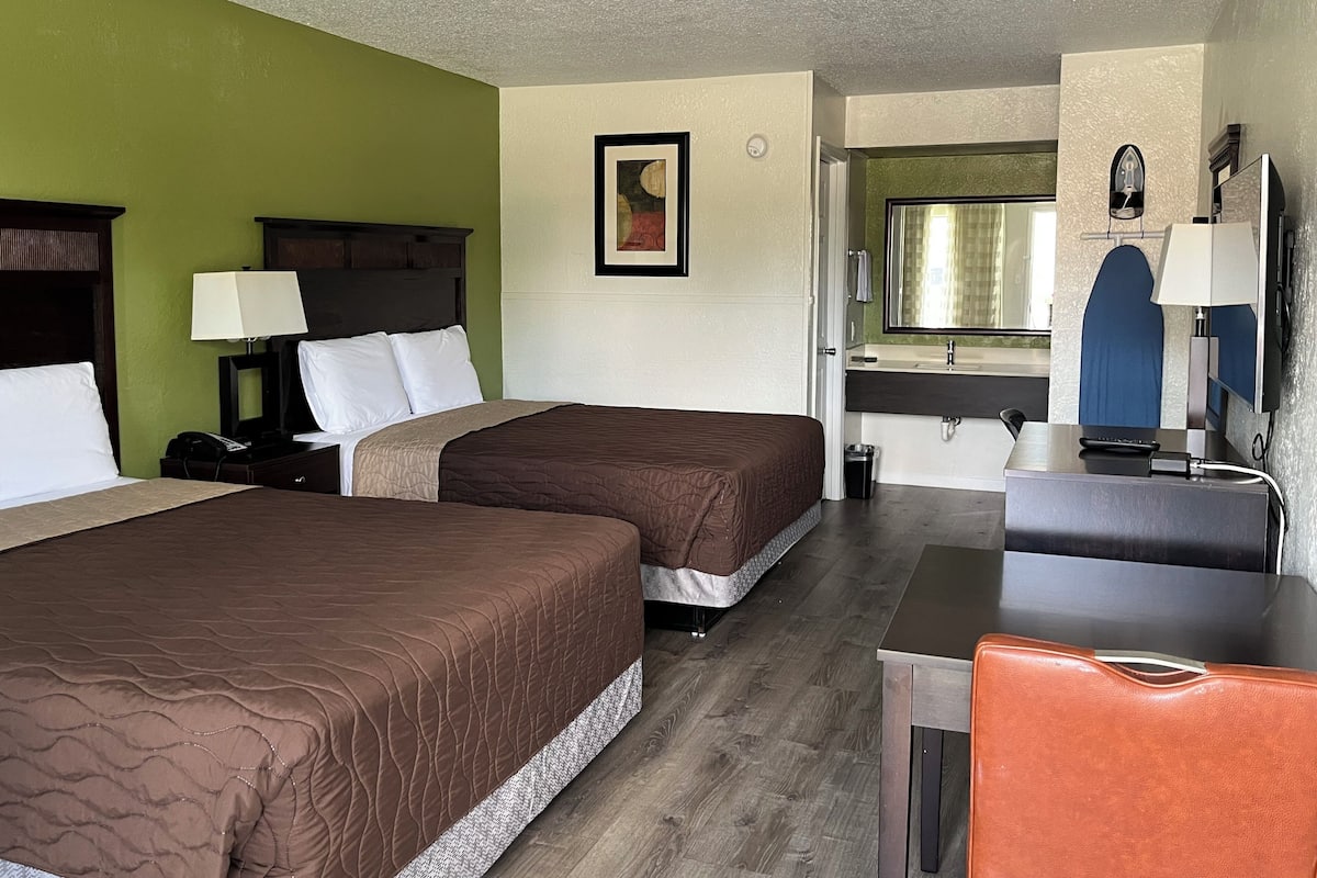 A hotel room with two beds, a desk, a chair, a mirror, and a bathroom visible through an open door.