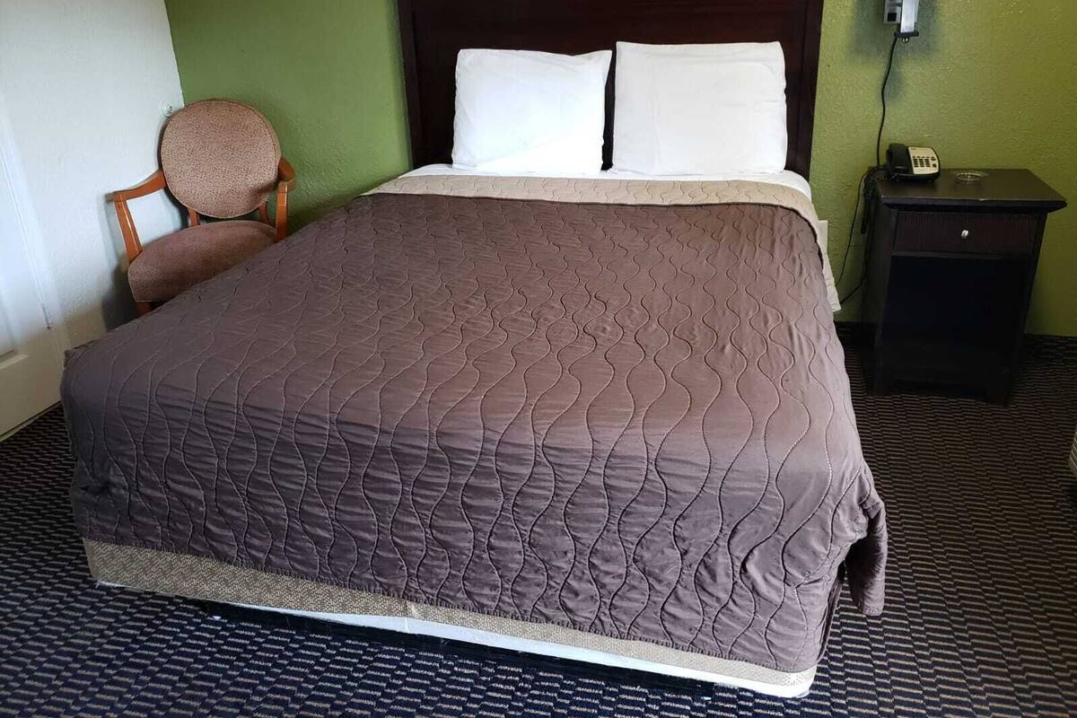 A hotel room with a large bed, a chair, a nightstand with a lamp, and a phone.