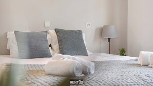 A neatly made bed with white and gray linens, a lamp, and a small plant on the side.