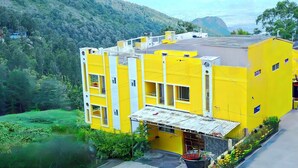A yellow building with multiple floors, surrounded by lush greenery and hills.