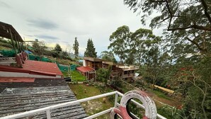 A view from a balcony with a metal railing, a tire sculpture, and a building with a red roof in the background. - Misty 7-Bedroom Hill Retreat in Kodaikanal–Perfect for groups & Artists (Kodaikanal)