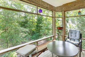 A wooden deck with a table and a cushioned chair, overlooking a forested area through large windows. - Fish On-Site: Rappahannock River Gem! (Urbanna)