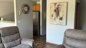 Uma sala de estar com dois sofás cinza, piso de madeira, cozinha ao fundo e um quadro na parede.