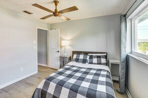 Une chambre à coucher comprenant un lit, un ventilateur de plafond, une table de chevet avec une lampe, une fenêtre avec des rideaux et une porte.