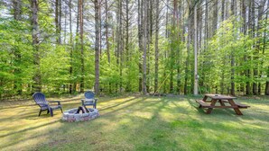 Un área de picnic con un brasero, dos sillas Adirondack y una mesa de picnic en un entorno boscoso.