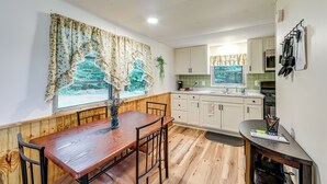 Una cocina con suelo de madera, armarios blancos, una mesa de comedor con sillas y una ventana con cortinas florales.