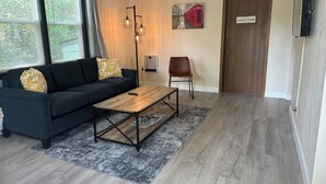 A modern living room with a dark grey sofa, a wooden coffee table, and a red chair. A painting of a red telephone booth is on the wall, and a sign reads "Laundry Room".