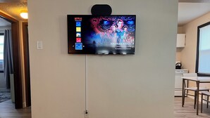 A flat-screen TV mounted on a wall in a room with wooden flooring and a kitchen area visible to the right.