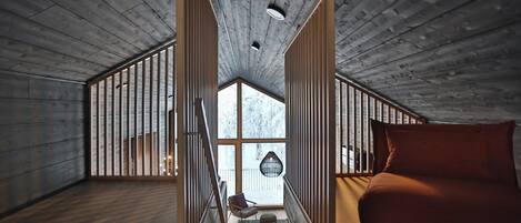 A high-ceilinged room with wooden beams, a bed, a chair, and a view through a large window.