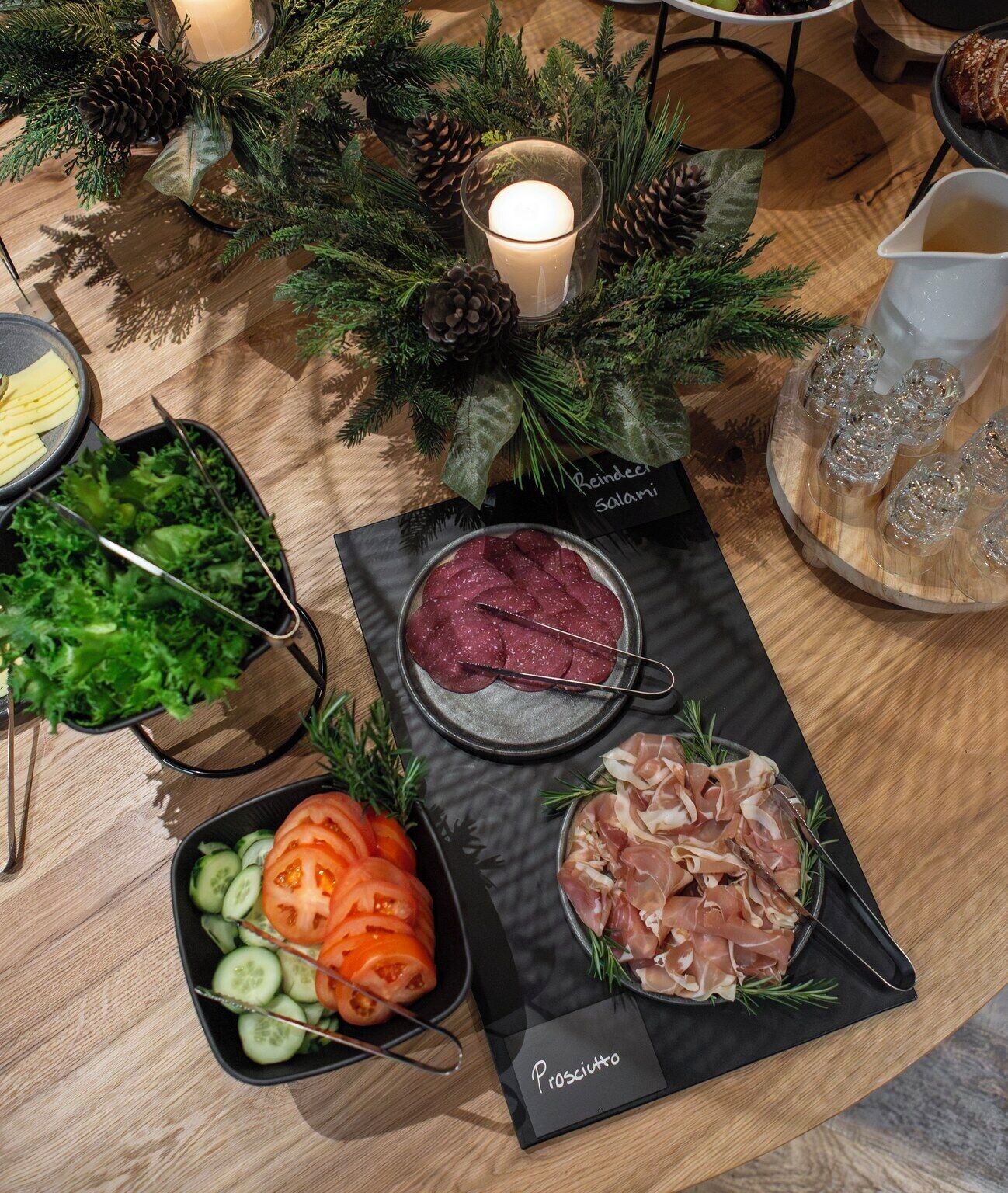 A table with a variety of food items including Prosciutto, Reindeer Salami, and a dish with tomatoes and cucumbers.