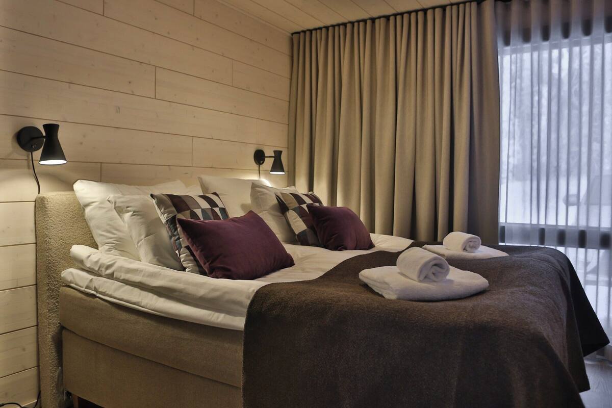 A hotel room with a bed, two wall-mounted lamps, a window with curtains, and towels on the bed.