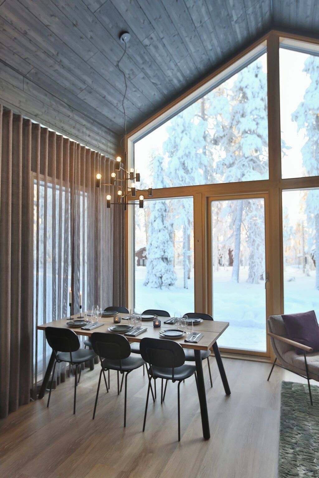 A dining area with a wooden table set for six, large windows with a snowy view, and a modern ceiling design.