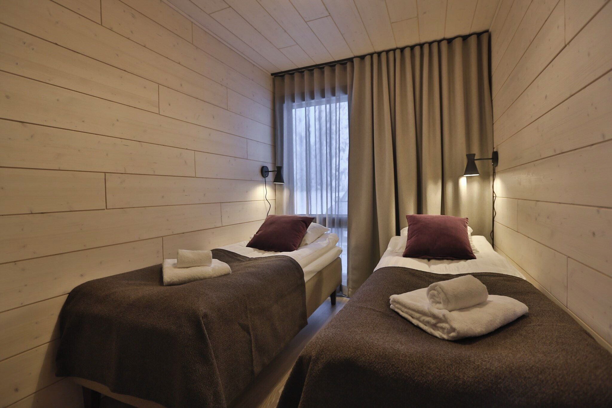 Une chambre avec deux lits, chacun recouvert d’une couverture marron et doté d’un oreiller, une fenêtre avec des rideaux et des lampes fixées au mur.