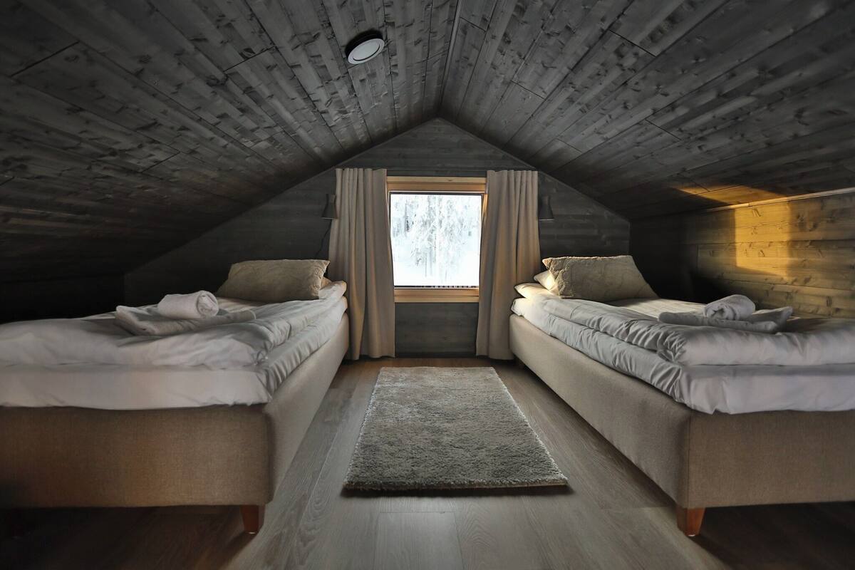 A cozy attic bedroom with two beds, a large window, and a wooden ceiling.