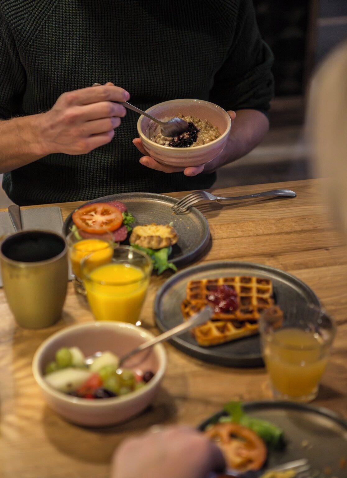 En frukostuppläggning med en skål med flingor, en tallrik med en grillad tomat, en våffla och en skål med sallad.