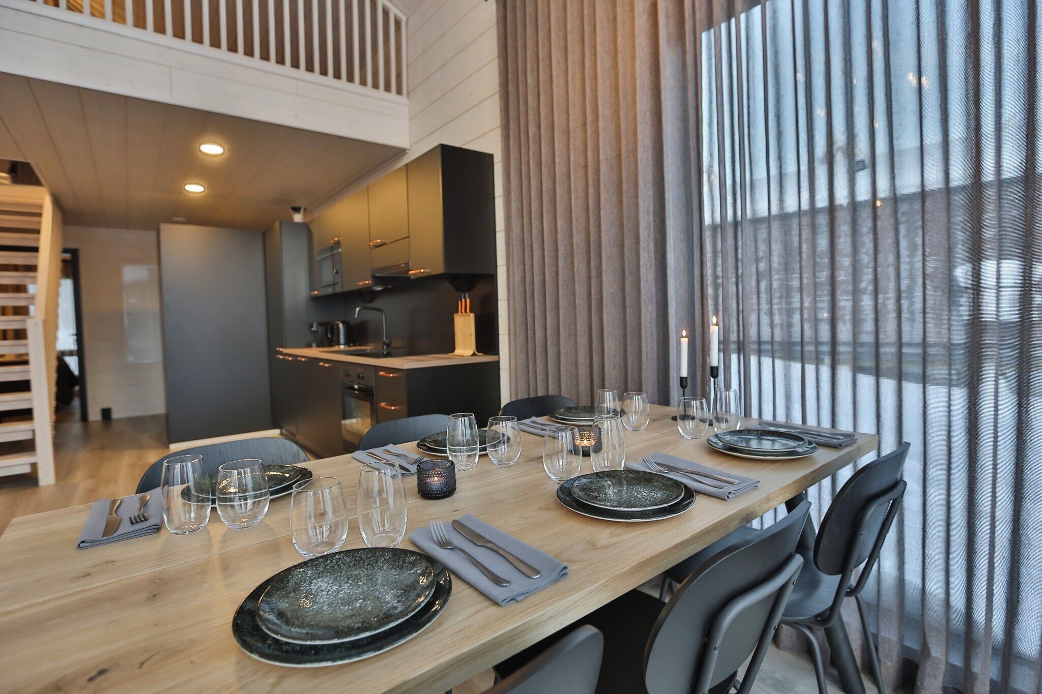 Un comedor moderno con una mesa larga dispuesta para seis personas, una cocina al fondo y ventanas amplias con cortinas.