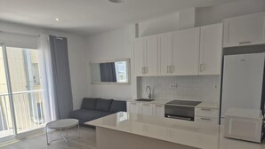 Une cuisine moderne avec des armoires blanches, une cuisinière encastrée et un réfrigérateur. On y trouve un petit îlot central avec un évier, ainsi qu’un coin repas avec une table et des chaises blanches.