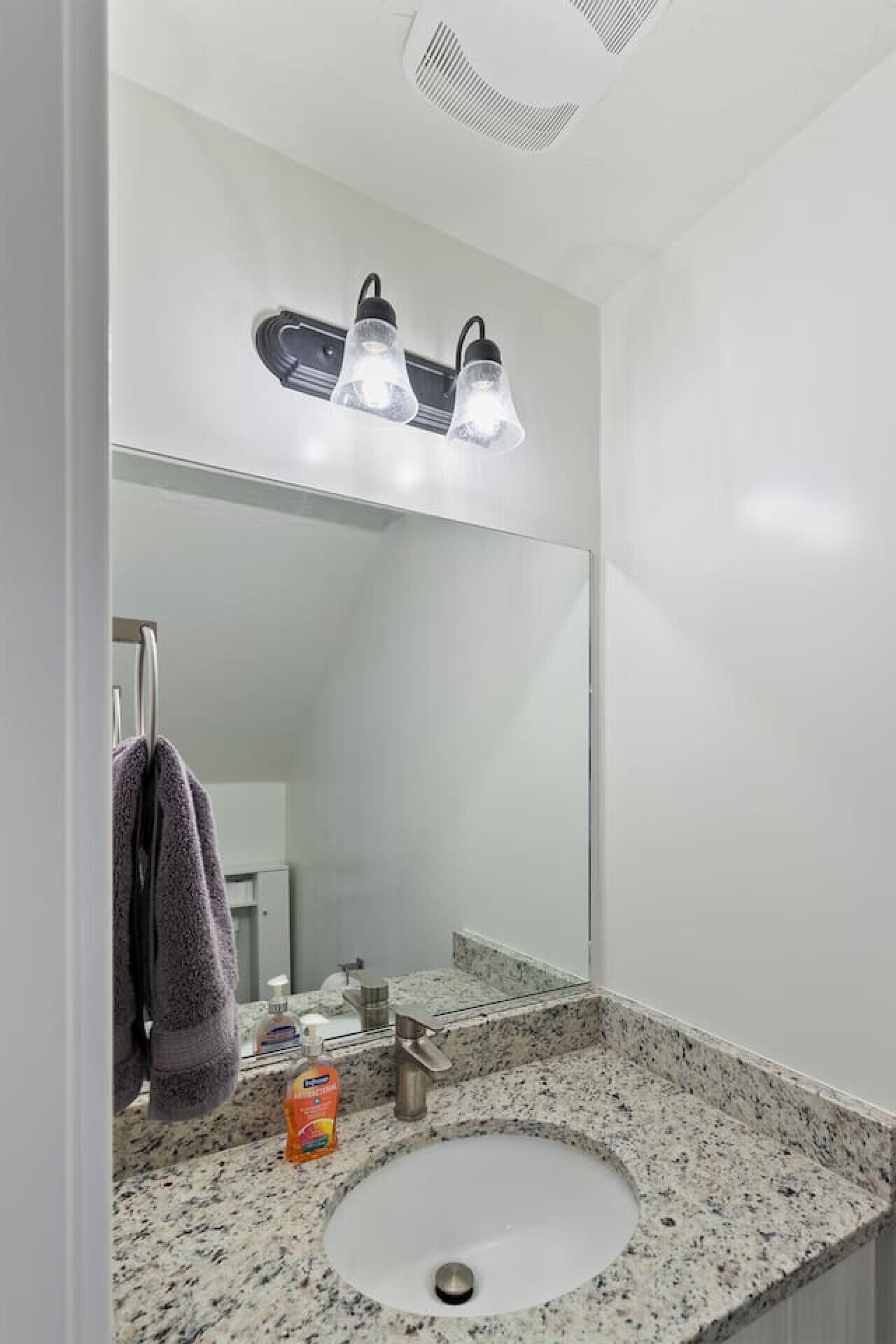 Ein Badezimmer mit einem weißen Waschbecken, einer Granit-Arbeitsplatte und einem Spiegel darüber. Zwei Leuchten sind über dem Spiegel angebracht.