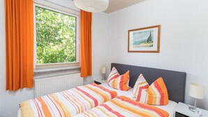 A double bed with striped bedding, a nightstand with a lamp, a framed picture on the wall, and a window with curtains. - Holiday Home 'Krohse' with Shared Garden and Wi-Fi (Potsdam)