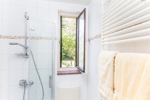 A bathroom with a glass shower enclosure, a window with a view of greenery, and a towel rack with a towel.