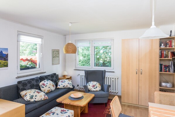 Una sala de estar con sofá, sillón, mesa, estantería y ventanas amplias.