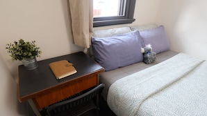 Ein kleines, aufgeräumtes Schlafzimmer mit einem Bett, einem Schreibtisch mit Notizbuch, einer Topfpflanze und einem Fenster mit Vorhängen.
