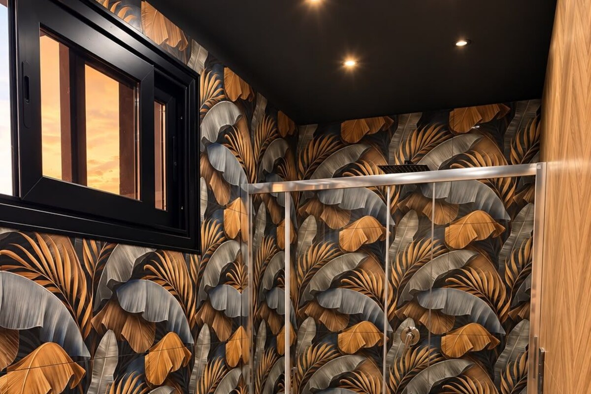 A modern bathroom with a leaf-patterned wallpaper, a wooden floor, a white sink, and a glass shower enclosure.
