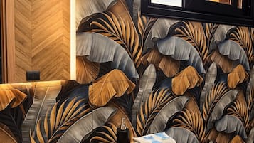 Een kamer met een houten visgraatvloer, een bank met behangpapier met bladmotieven, een raam met jaloezieën en een tafeltje met een handdoek en een fles.