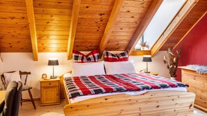Ett sovrum i loftstil med ett sluttande tak, synliga träbjälkar, en säng med ett rutigt överkast, två sängbord med lampor och en röd accentvägg.