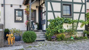 Ett traditionellt hus med korsvirkesfasad, en trappa i sten, en hundskulptur och diverse krukväxter.
