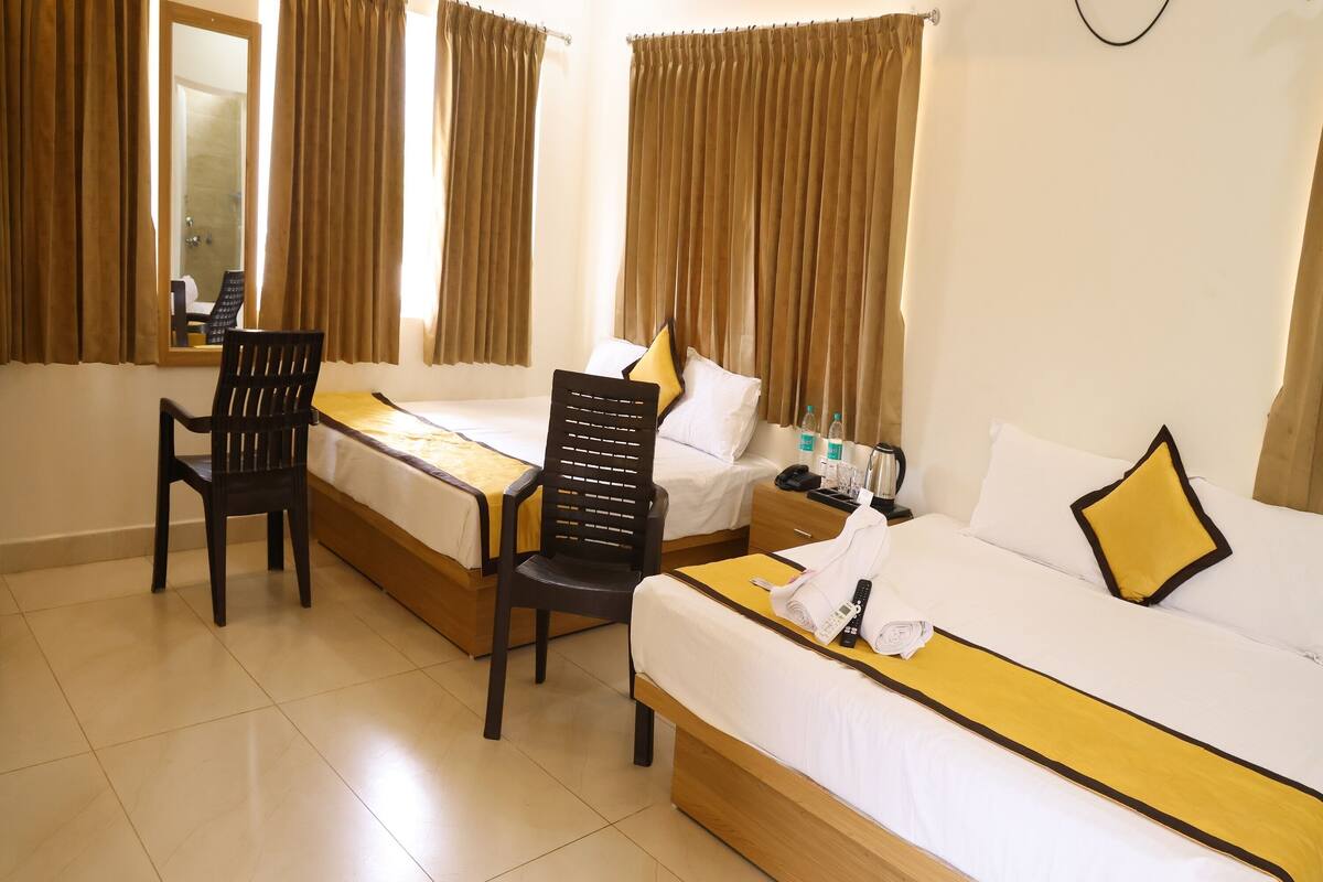 A hotel room with two beds, a desk, and a chair. There is a window with curtains, an air conditioning unit, and a mirror on the wall.