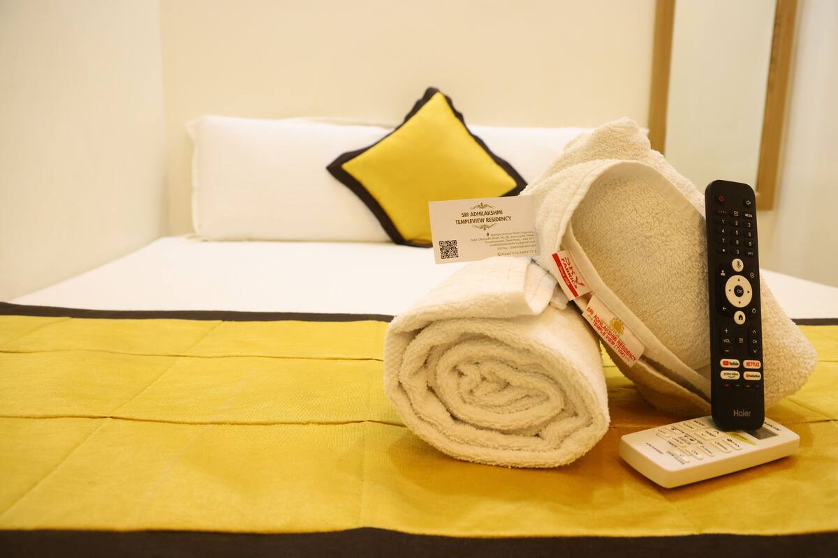 A neatly made bed with a white and yellow bedspread, a rolled white towel, a remote control, and a card on the bed.