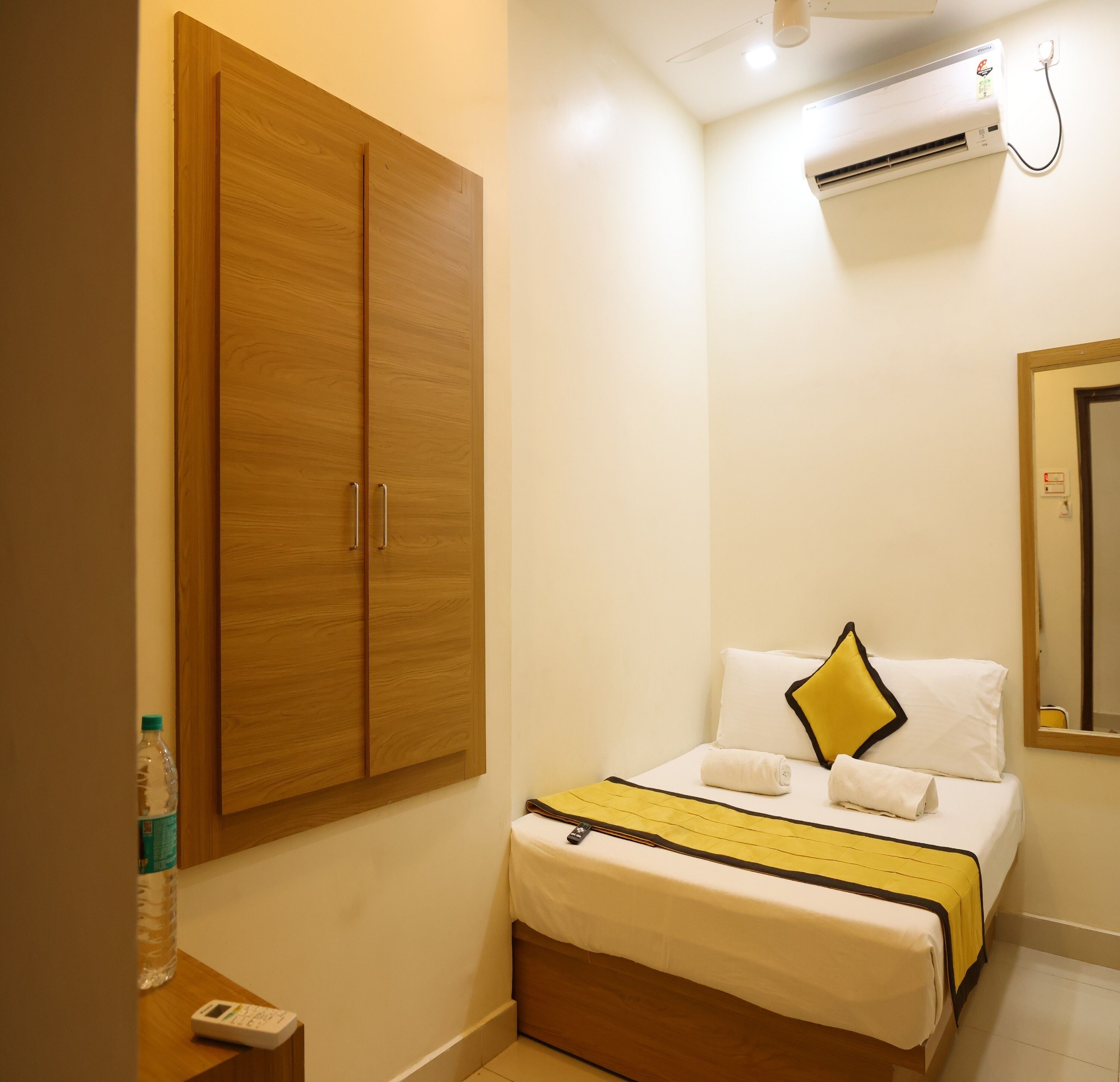 A hotel room with a wooden wardrobe, a bed with white bedding and a yellow accent pillow, an air conditioning unit, and a bottle of water on the nightstand.