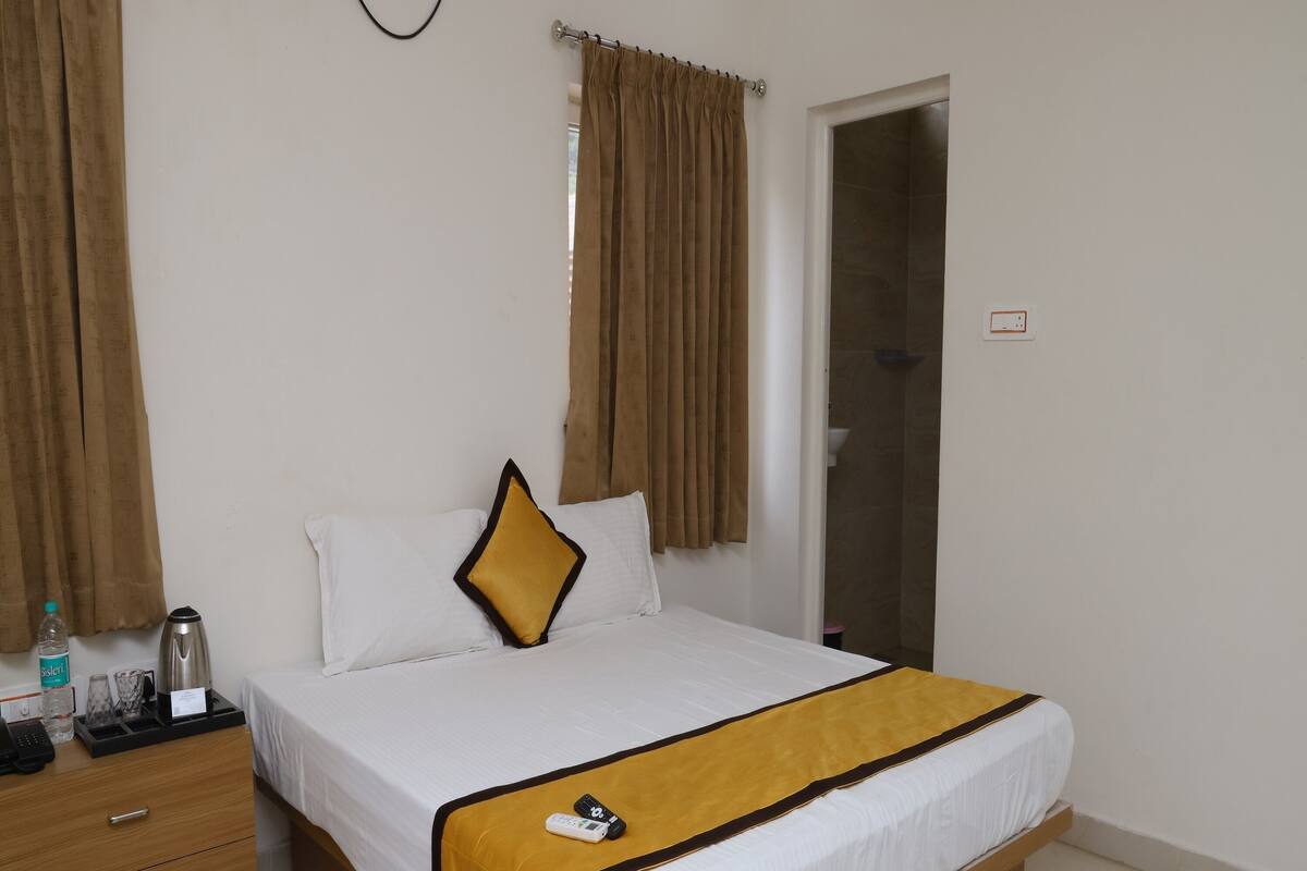 A hotel room with a bed, a bedside table with a phone and a bottle, a wooden nightstand, a bathroom with a shower, and a door with a curtain.
